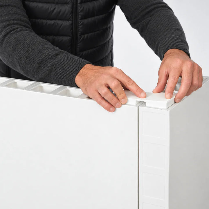 A close-up of a person's hands installing a white plastic top panel onto a white cabinet or refrigerator, wearing a black quilted jacket.