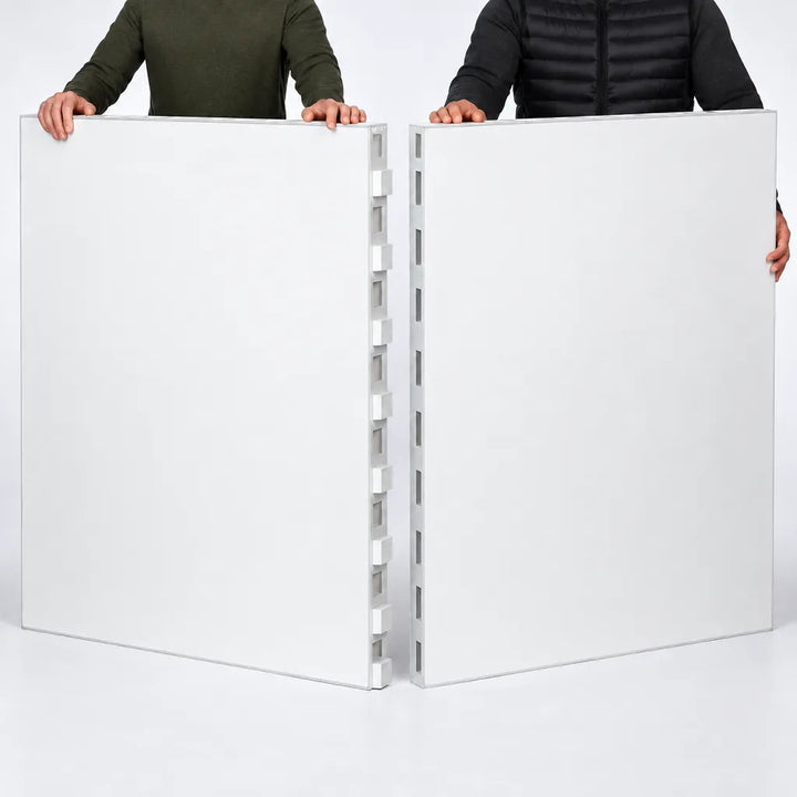 Two people holding up two large white interlocking panel pieces, likely modular wall or ceiling tiles, against a clean white studio background.
