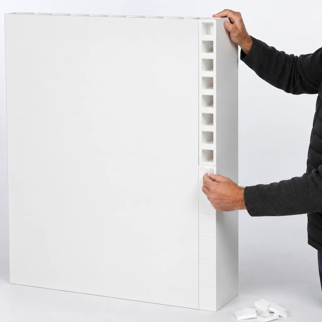 A person installing a white modular wall panel with a vertical row of square cutouts, shown against a clean white studio background.